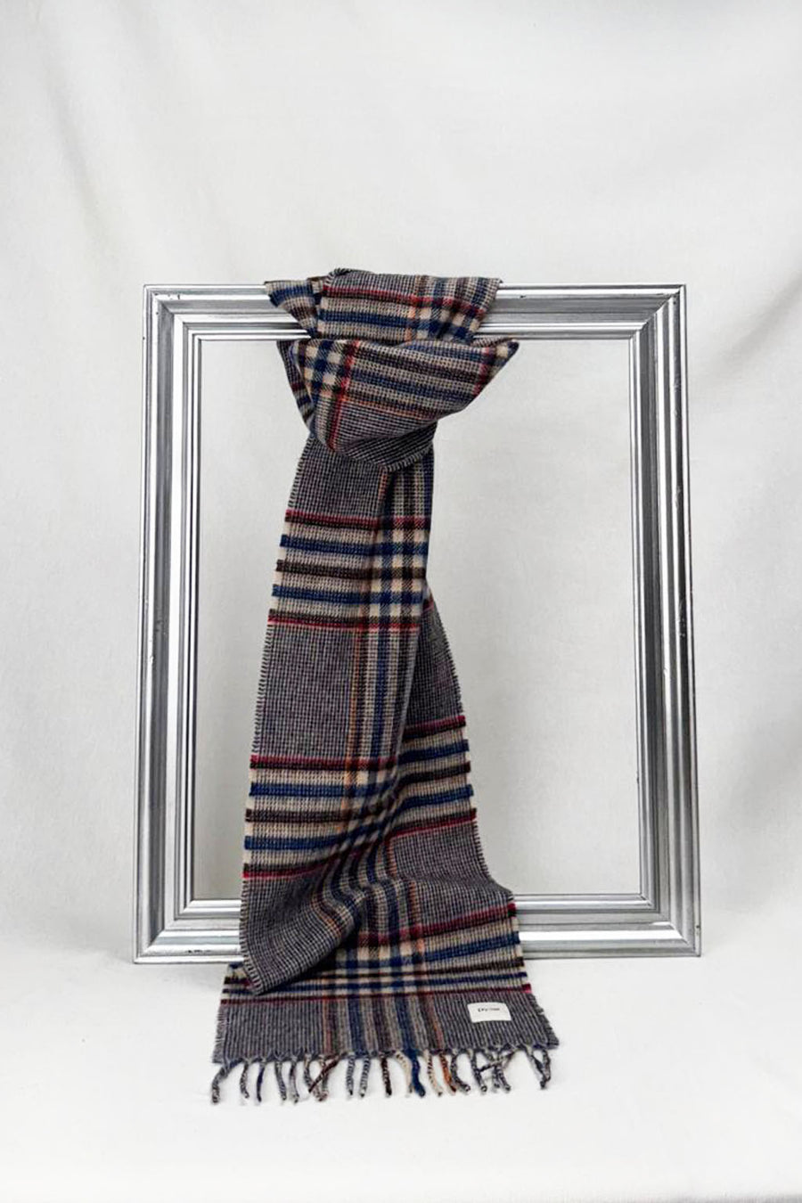 A neatly draped grey plaid wool scarf featuring navy blue, beige, and red stripes hangs over a silver frame. The soft fabric and refined pattern highlight sophistication and warmth, making it a perfect winter accessory or thoughtful gift for her with delivery across Malta.