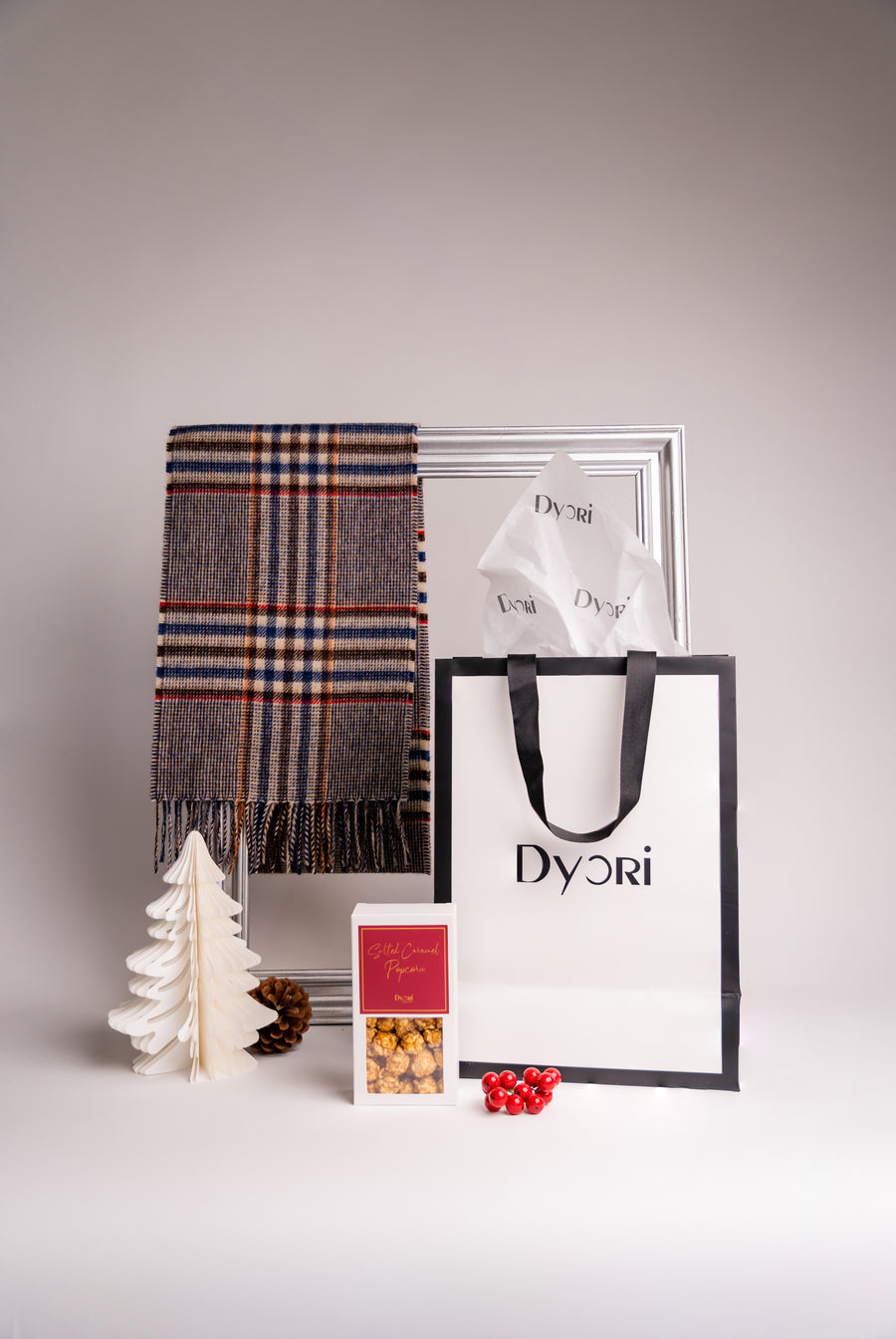 A timeless merino wool scarf in brown and navy plaid tones, styled with a Dyori gift bag, golden popcorn box, pine cone, and festive berries. The image reflects elegance and premium craftsmanship — ideal for holiday gifting.