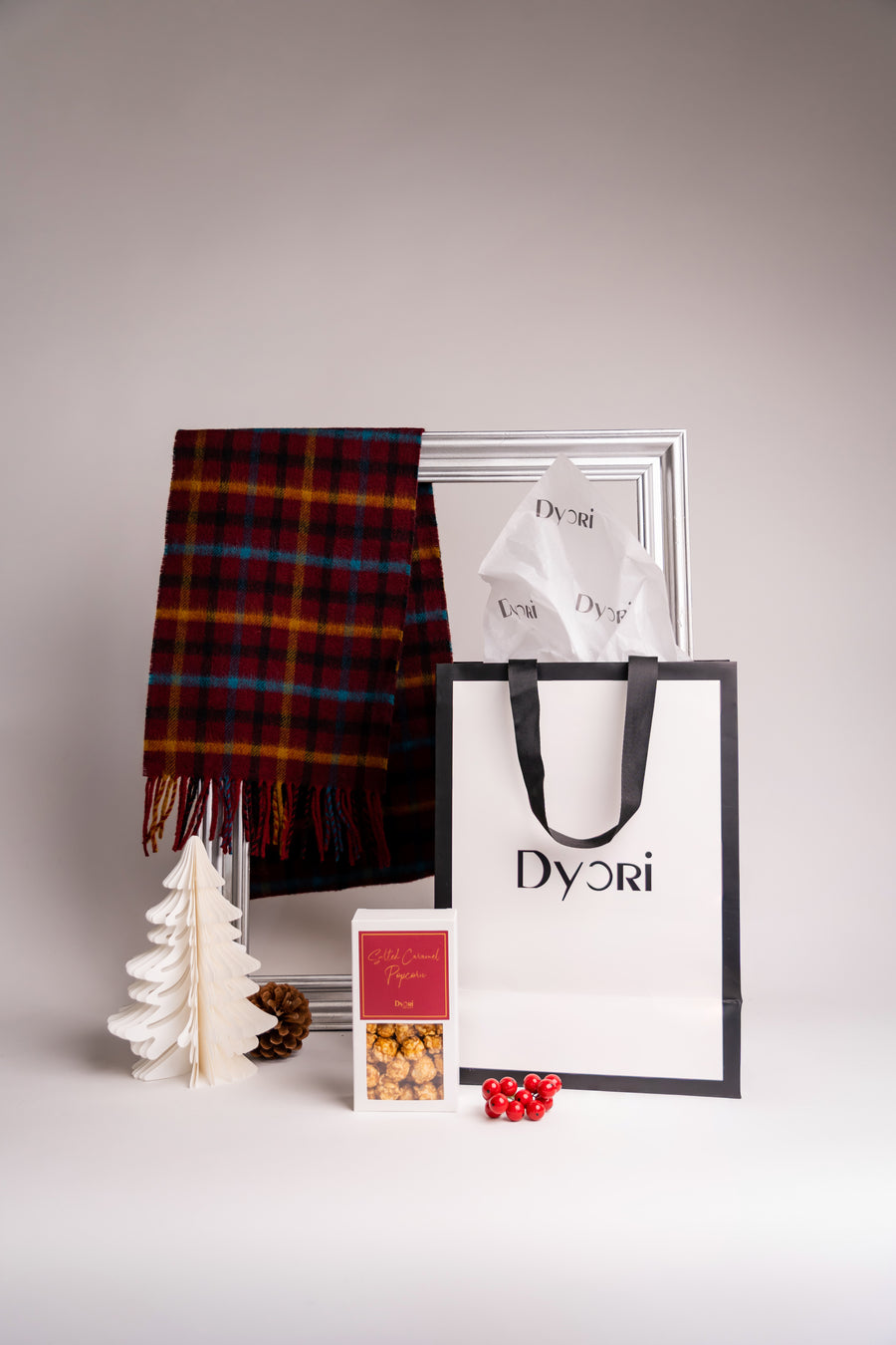 A soft merino wool scarf in deep red with striking mustard and blue plaid detailing. Presented beside a Dyori gift bag, caramel popcorn, and holiday decor, the image evokes warmth and sophistication for the winter season.