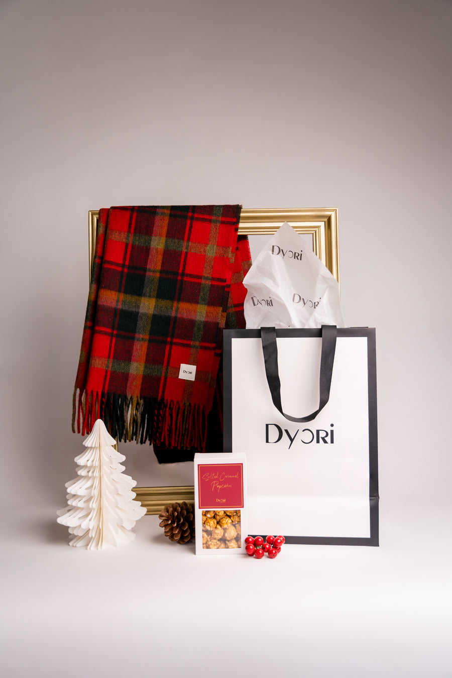 A festive Dyori red and green check-pattern cotton scarf styled beside a white Christmas tree, pinecone, and luxury gift bag—perfect for cozy holiday gifting.