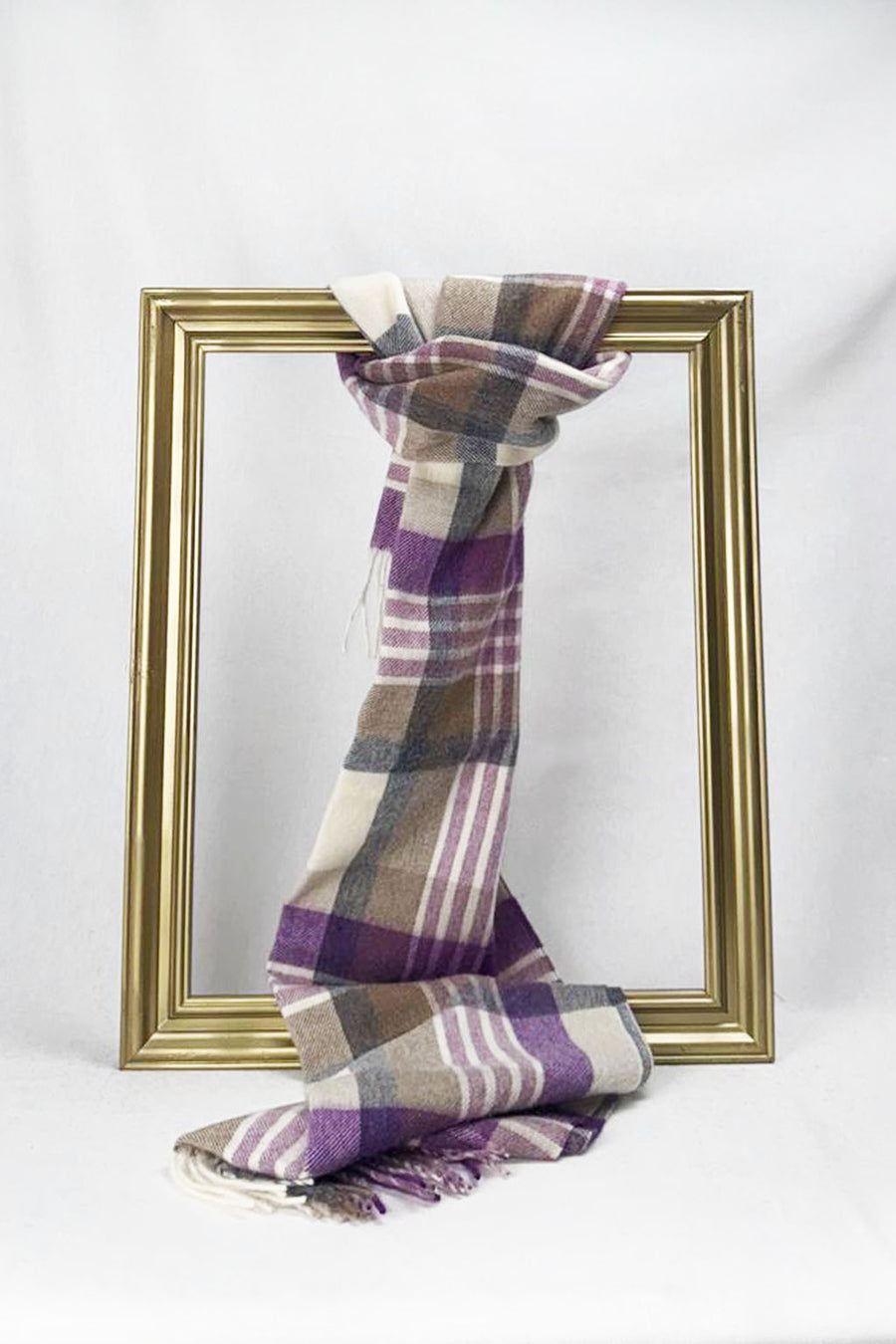 A beautifully folded plaid wool scarf in purple, tan, grey, and cream hangs over a gold picture frame on a clean white backdrop. The minimalist setup showcases the scarf’s premium quality and timeless checkered pattern, perfect as a thoughtful gift for her. Enjoy convenient delivery across Malta.