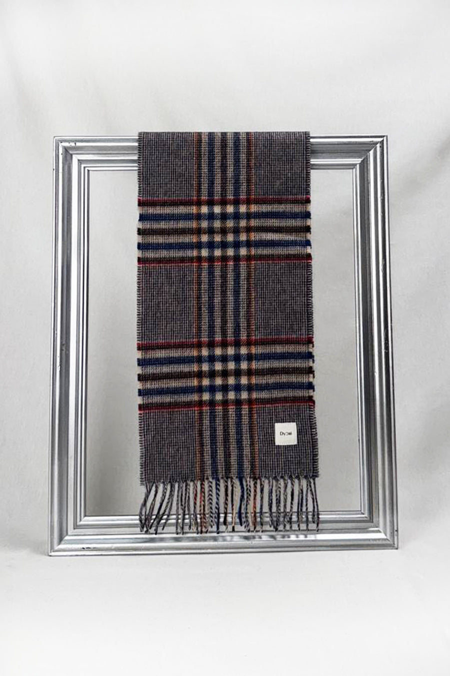 A stylish grey wool scarf featuring a check pattern with navy blue, beige, and red stripes is gracefully looped through a silver frame. Displayed against a clean white background, this soft plaid scarf embodies timeless winter fashion — a warm and elegant gift for her. Available with delivery across Malta.