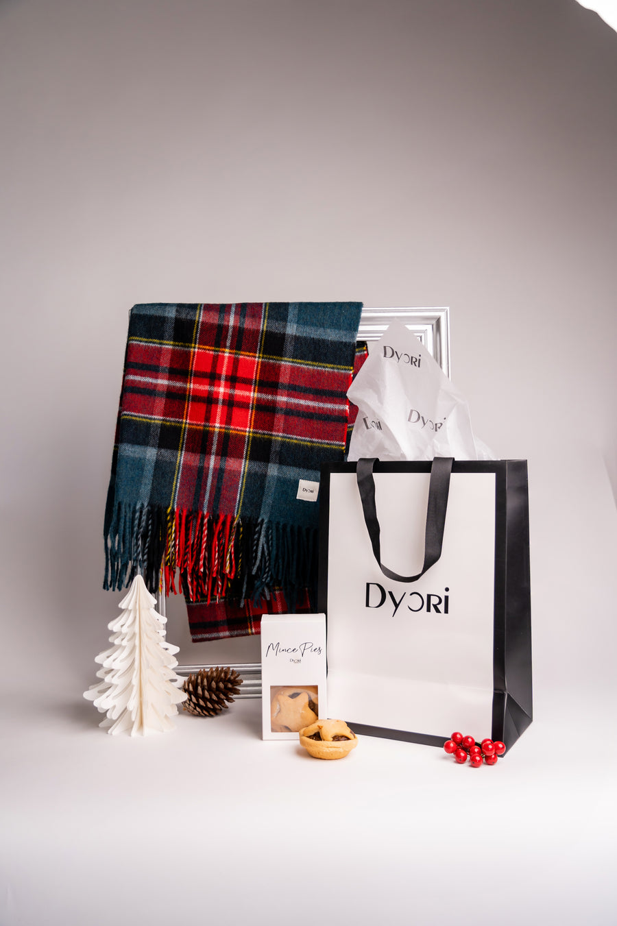 Dyori’s bold red and teal wool plaid scarf displayed in a joyful Christmas setup, complete with pinecones, berries, and Dyori’s elegant packaging
